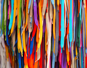 A vibrant display of multicolored shoelaces hanging in rows, featuring a wide array of colors like blue, red, yellow, green, orange, and purple with various textures and patterns.
