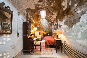 Al Capone's prison cell at Eastern State Penitentiary, featuring a bed with a red blanket, antique wooden furniture, a vintage lamp, a small writing desk, and peeling walls with exposed plaster, creating a historic and somewhat eerie atmosphere.