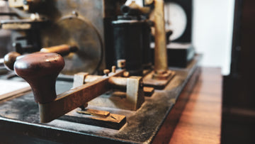 An antique Morse code telegraph machine with a prominent telegraph key in the foreground, and various mechanical parts out of focus in the background, on a wooden surface.